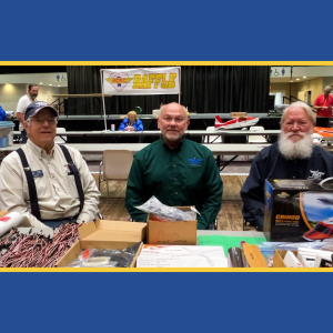 Three men seated at a table with model airplane kits, indoors at an event.