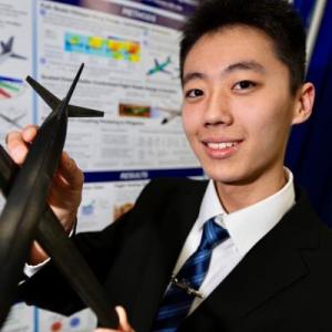 Young man in a suit holds a model aircraft, smiling, in front of a scientific poster.