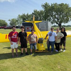 Seven people stand in front of a small, yellow airplane on a grassy field.