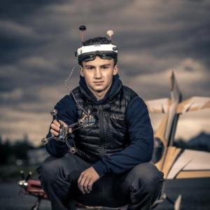 Drone racer holding quadcopter, wearing VR goggles, with blurred landscape backdrop.