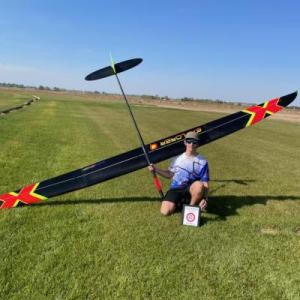 Person kneeling on grass with a large model glider against a clear blue sky.