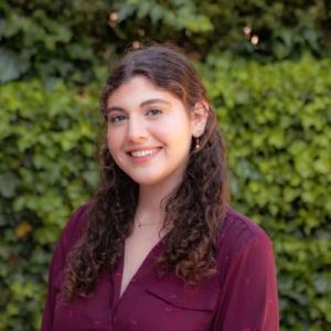 Smiling person with curly hair in a maroon shirt, greenery in the background.