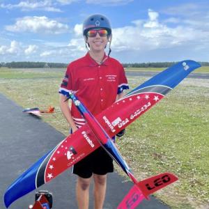 Teen holding a red and blue model airplane, wearing a helmet and red shirt outside.