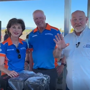 Man greets camera beside two smiling individuals at an outdoor event.