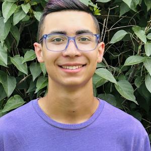 Young man in glasses and purple shirt smiling, standing in front of leafy background.