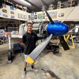 Young person standing near a disassembled small aircraft engine in a workshop.
