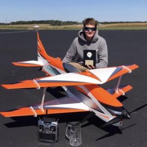 Young man kneeling by a large orange model plane, holding a plaque, with remote control nearby.
