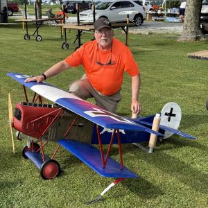 Man kneeling on grass next to a large red-and-blue model biplane.