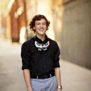 Young person with curly hair smiling, wearing a black shirt, standing outdoors.