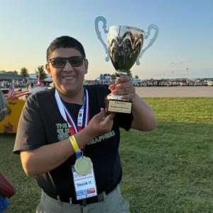 Person smiling and holding a trophy outdoors on a sunny day.