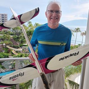 Man smiling, holding two model gliders on a balcony in a tropical setting.