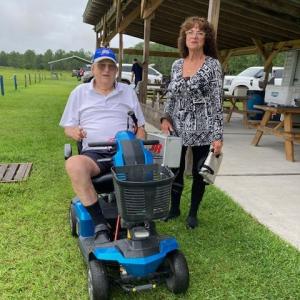 Man on mobility scooter with woman standing nearby under a wooden pavilion.