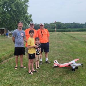 Four people standing on a grassy field beside a model airplane.