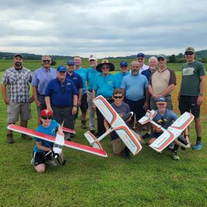 Group of people outdoors holding model planes on a grassy field.