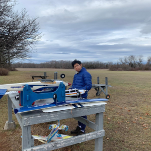 Person in a blue jacket assembling a model airplane on a wooden table outdoors.