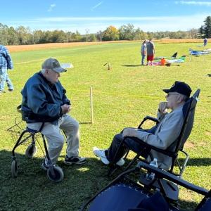 Two elderly men sitting outdoors on a grassy field, with model planes in the background.