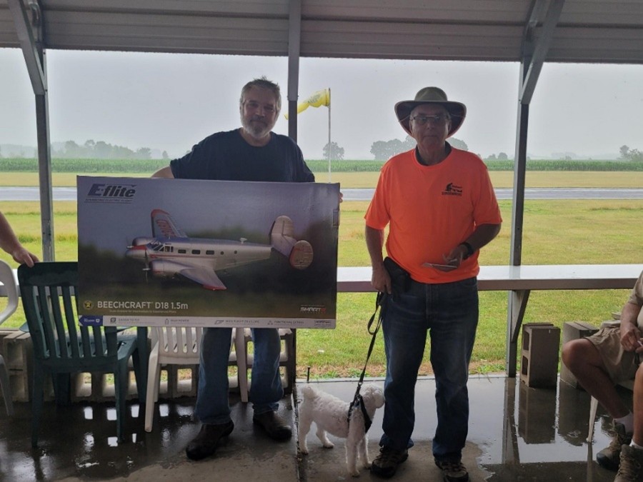 Two men holding a model airplane box under a canopy on a rainy day.