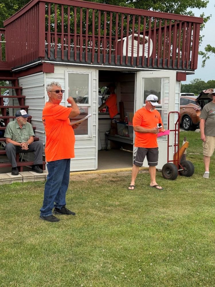 Man in orange shirt standing on grass, others seated nearby, next to a small white building.