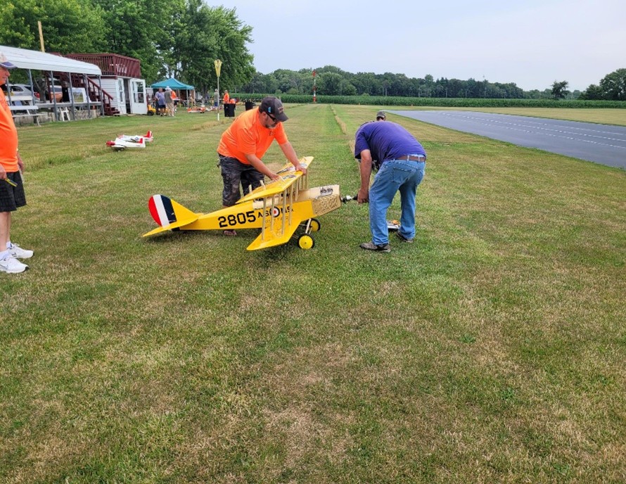 Two people adjust a small yellow model aircraft on grass near a runway.