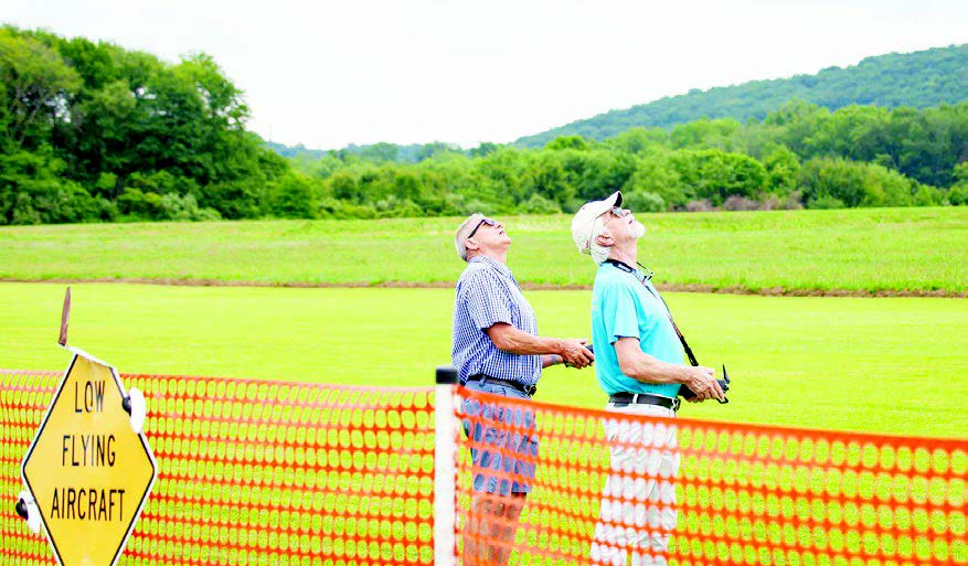 Two men look up, controlling a model aircraft near a grassy field.