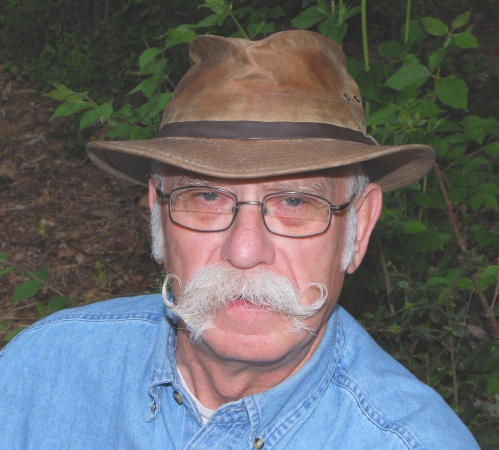 Elderly man with a mustache and glasses wearing a brown hat and blue denim shirt.