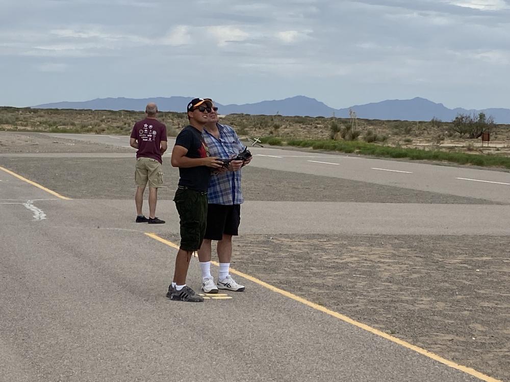 Two people standing on a remote tarmac, one holding a remote control, looking skyward.
