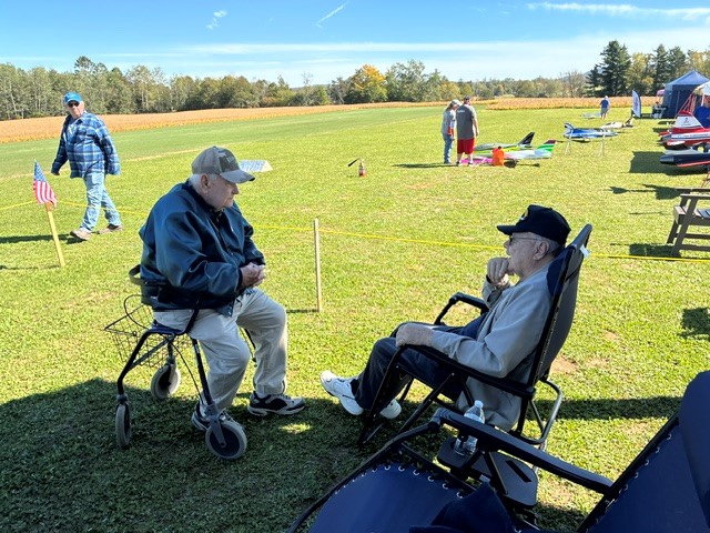 Two elderly men in conversation, seated on grass on a sunny day.