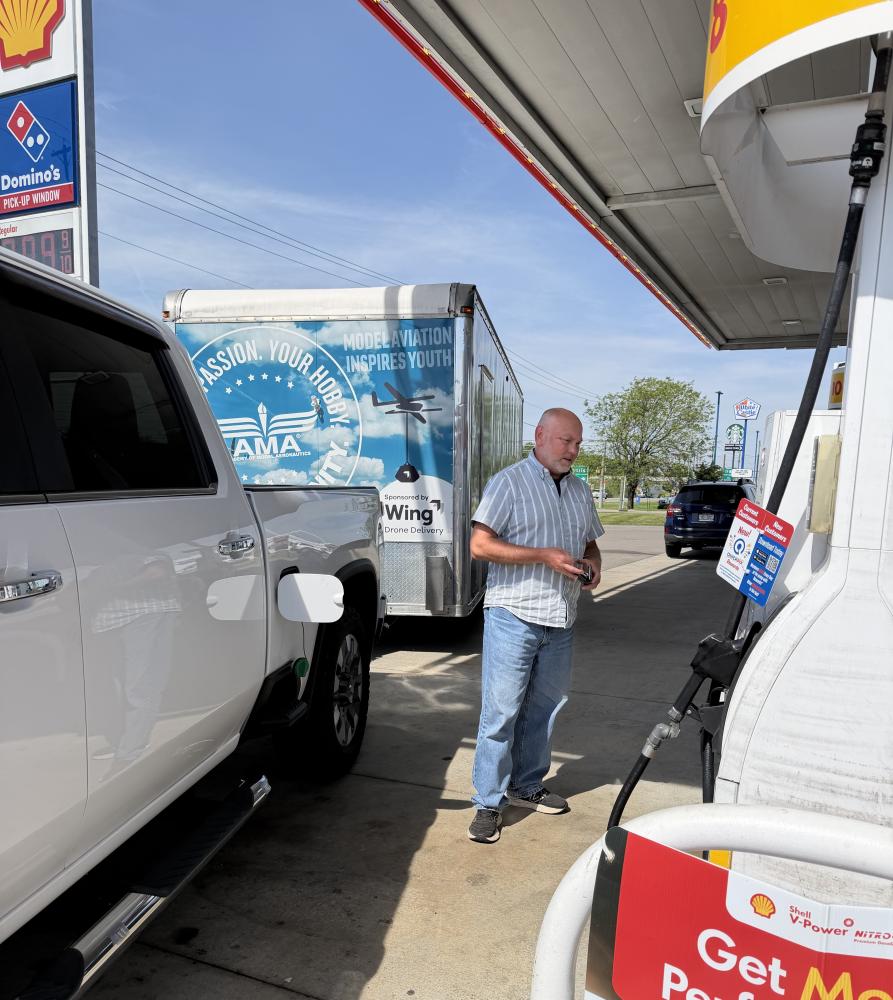 Man standing at a gas station, with a white truck and a trailer in the background.