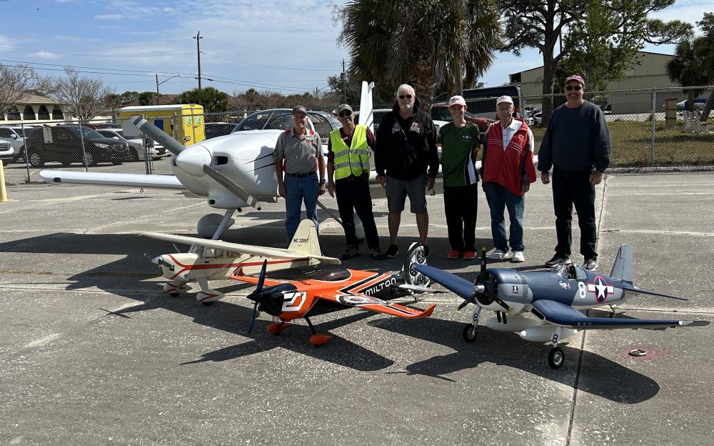 Seven people standing by model airplanes on a sunny day.