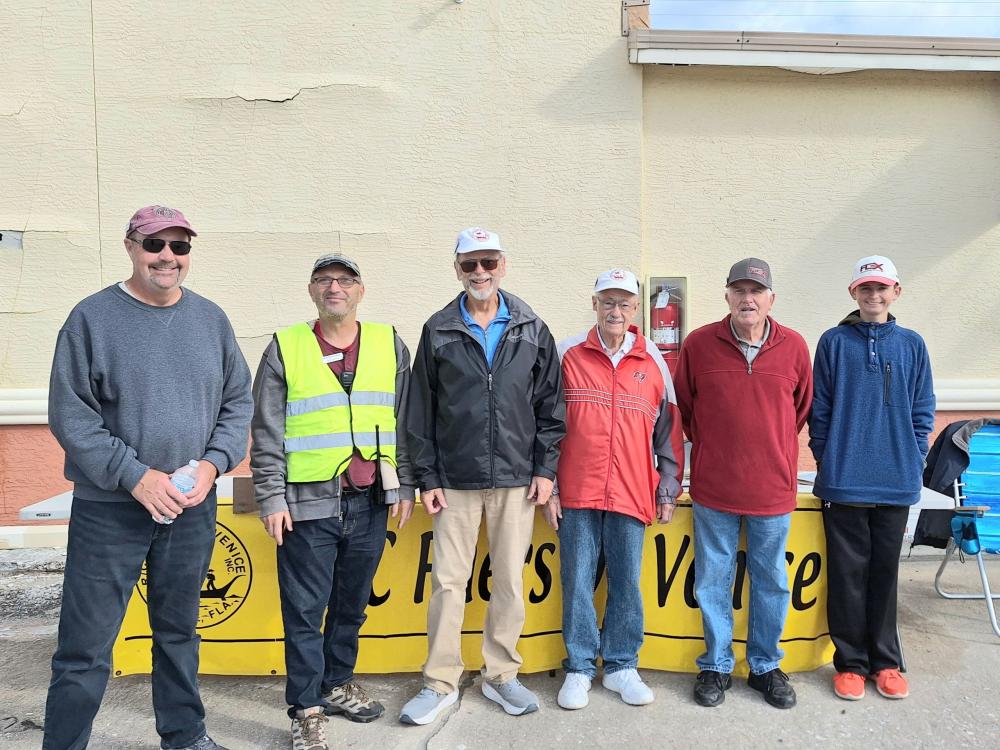 Six men standing in front of a yellow table with a wall behind them, smiling at the camera.