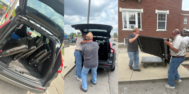 Three men unloading metal racks from a car onto a dolly outside a brick building.